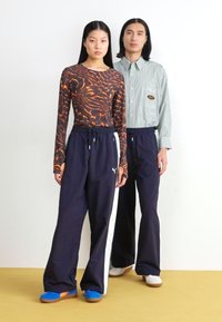 Μπλούζα με μακρύ μανίκι και αφηρημένο πορτοκαλί σχέδιο, συνδυασμένη με ναυτικές παντελόνες που έχουν πλευρική γραμμή. Μοντέλο που φοράει μπλε παπούτσια και λευκά αθλητικά.