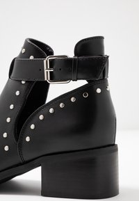 Botines negros de piel con tacón grueso, que cuentan con un cierre lateral con hebilla y acentos de tachuelas plateadas a lo largo de la abertura y los lados.