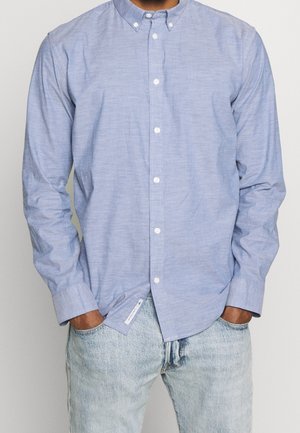 Mand iført lyseblå langærmet skjorte med knapper og lyse cowboybukser, hænder i lommerne, stående mod en ensfarvet baggrund.