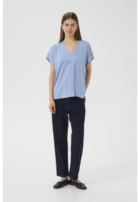 Blusa azzurro chiaro a maniche corte con scollatura a V, tessuto morbido, vestibilità rilassata, abbinata a pantaloni dritti in denim scuro e scarpe nere con suola piatta.