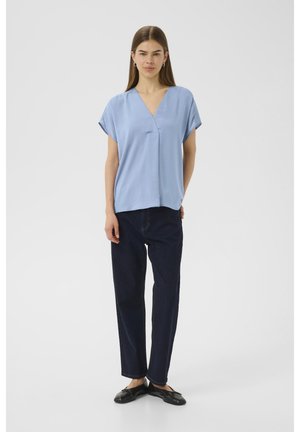 Blusa azzurro chiaro a maniche corte con scollatura a V, tessuto morbido, vestibilità rilassata, abbinata a pantaloni dritti in denim scuro e scarpe nere con suola piatta.