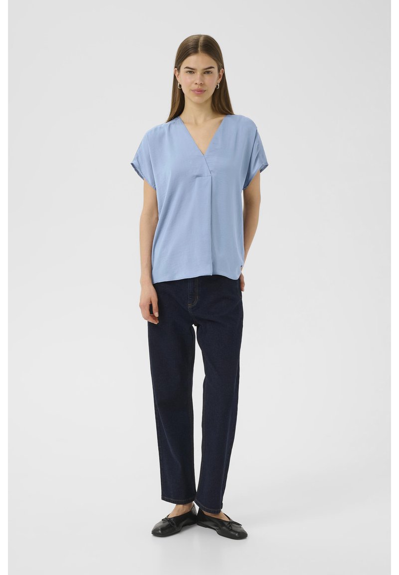 Blusa azzurro chiaro a maniche corte con scollatura a V, tessuto morbido, vestibilità rilassata, abbinata a pantaloni dritti in denim scuro e scarpe nere con suola piatta.