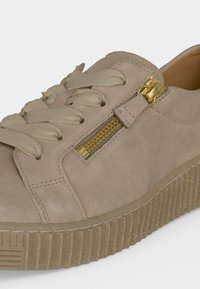 Baskets en daim beige avec une semelle épaisse à relief, de larges lacets et un détail de fermeture éclair dorée sur le côté près des lacets.