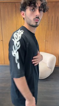 Camisa T-shirt oversized preta com design gráfico branco ao longo da manga esquerda. Apresenta um corte relaxado e uma textura de tecido suave.
