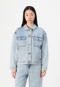 Chaqueta de mezclilla azul claro con botones en el frente, dos bolsillos de parche y un corte relajado. Textura suave con un diseño informal y costuras visibles.