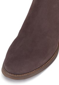 Brązowy zamszowy botek za kostkę z zaostrzonym noskiem i widocznym przeszyciem wzdłuż podeszwy. Gładka tekstura i prosty, czysty design.