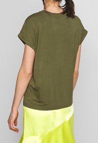 Olivově zelené bavlněné tričko s krátkými ohrnutými rukávy, volným střihem a zakřiveným spodním lemem. Nosené s jasně žlutou saténovou sukní.