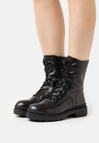 Stivali alla caviglia in pelle nera con lacci frontali, punta rotonda e suola in gomma testurizzata. Presentano occhielli metallici e una zip laterale per una facile vestibilità.