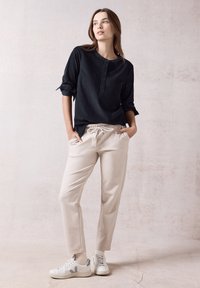 Camicia nera con bottoni e maniche arrotolate, pantaloni beige con cordino e sneakers bianche. Tessuto morbido e vestibilità rilassata.