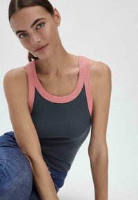 Donkergrijs geribbeld tanktop met een roze geribbelde halslijn, met brede banden en een getailleerd silhouet. Gemonteerd door een model, zittend tegen een neutrale achtergrond.