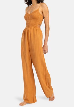Combinaison orange avec un corsage froncé, des bretelles fines et un pantalon large avec des rayures verticales discrètes et des découpes sur les côtés. Tissu texturé.
