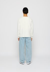 En person som har på seg en svart lue, en hvit langermet topp, lyseblå vide jeans og hvite sneakers, står med ryggen mot kameraet mot en ensfarget bakgrunn.