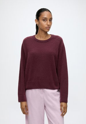 Vrouw staat frontaal, draagt een losse bordeaux trui en lichtroze broek tegen een effen lichte achtergrond, met het haar naar achteren gebonden.