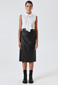 Vrouw in een zonder mouwen witte blouse met een decoratieve sluiting, zwarte leren rok tot de knie en zwarte veterschoenen, staand tegen een effen achtergrond.