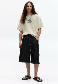 Béžové oversized tričko s grafickým potlačou, čierne džínsové šortky, čierne žabky a vrstvené strieborné a zlaté retiazky.