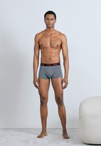 Gray cotton boxer shorts with a black waistband featuring red "HUGO" branding, displayed on a shirtless male model, standing against a light background.