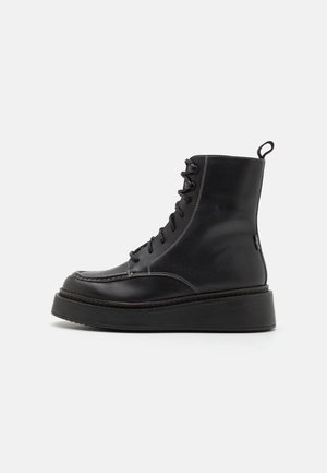 Botas de tobillo de cuero negro con punta redonda, suela gruesa y plataforma, y sistema de cordones. Presentan costuras en contraste y una asa en el talón.