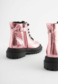 Bottines roses métalliques avec semelles noires épaisses, dotées d'un laçage à l'avant, d'une fermeture éclair sur le côté et d'une étoile en accent sur le talon. Fini texturé.