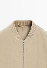 Beżowa kurtka z prążkowanym kołnierzem i srebrnym, centralnym zamkiem błyskawicznym, wykonana z teksturowanej tkaniny.