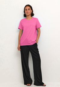 T-shirt en coton rose à manches courtes, col rond et coupe décontractée, associé à un pantalon large noir. Le mannequin se tient devant un fond clair.