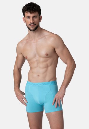 Uomo in forma con capelli corti e ricci che indossa slip azzurri chiari, in piedi contro uno sfondo chiaro e uniforme.