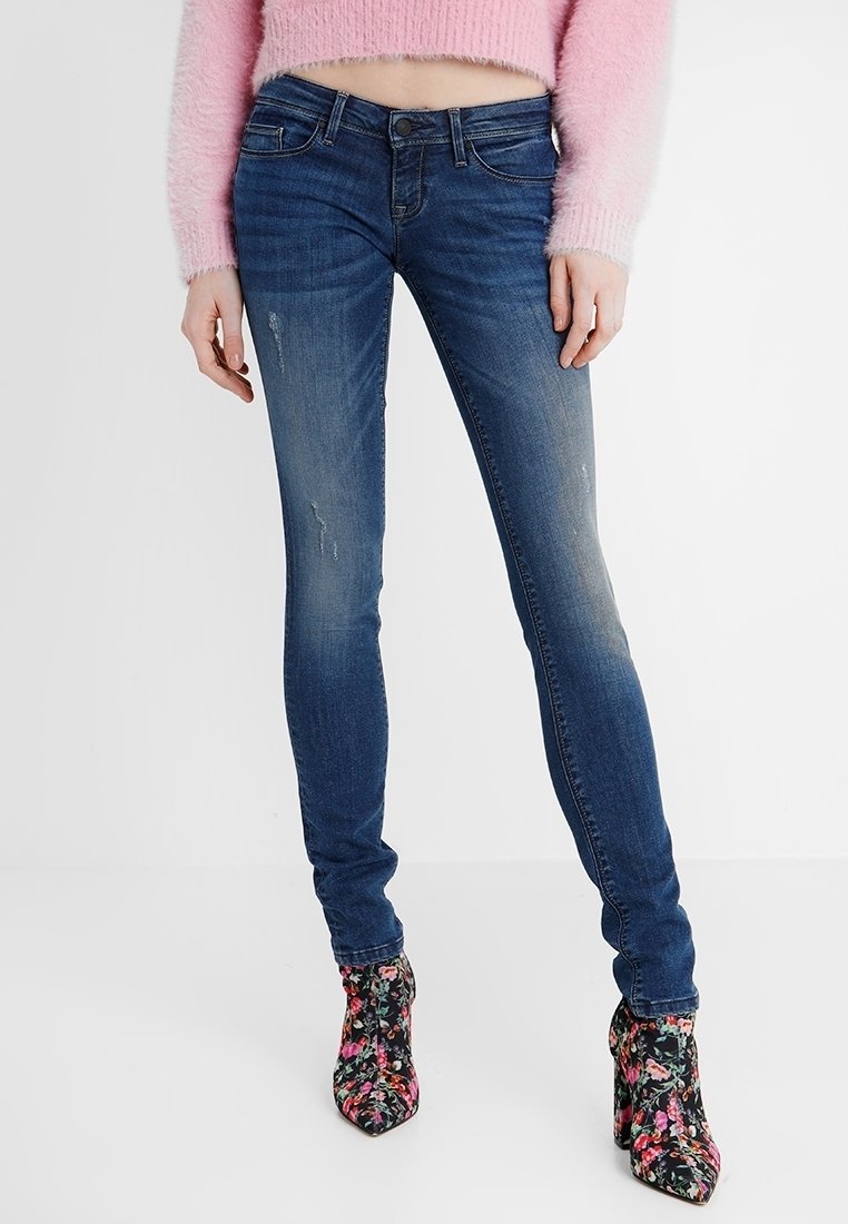 Schlanke blaue Jeans mit einem verblassten Finish, das leichte Abnutzungserscheinungen aufweist. Kombiniert mit blumendruckierten, hohen Stiefeletten.