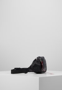 Mustavärinen nahkavyölaukku, jossa on teksturoitu pinta. Laukussa on etupuolella vetoketjullinen tasku ja säädettävä hihna. Vetoketjusta roikkuu punainen lapu.