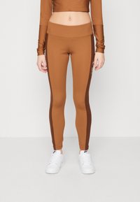 Hnědé sportovní legíny s vysokým pasem, které mají tmavě hnědé boční panely. Vypadávají v kombinaci s odpovídajícím dlouhým topem s krátkým rukávem a bílými teniskami.