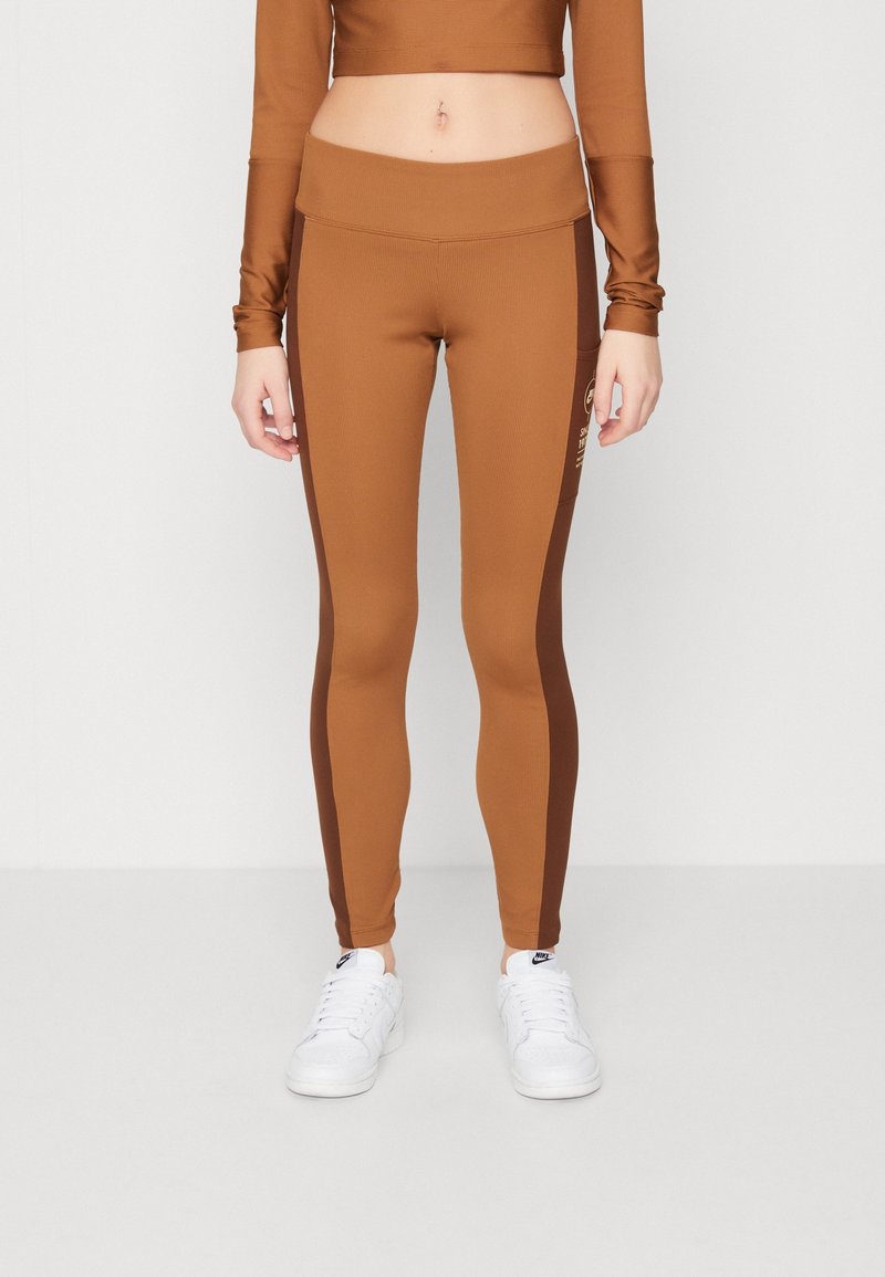 Hnědé sportovní legíny s vysokým pasem, které mají tmavě hnědé boční panely. Vypadávají v kombinaci s odpovídajícím dlouhým topem s krátkým rukávem a bílými teniskami.