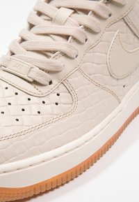 Närbild av en beige sneaker med krokodilmönstrad läder, perforerad tå, matchande snören och sula av gummiblandning.