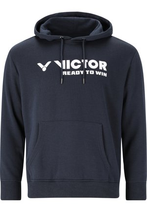 Lyshåret mand iført en mørkeblå hættetrøje med "VICTOR READY TO WIN" logo, stående mod en ensfarvet lys grå baggrund.