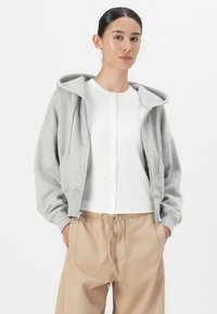 Sweat à zip gris avec une coupe standard, capuche à cordons et deux poches avant, porté sur une chemise blanche à boutons et un pantalon beige.