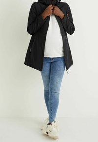 Musta kevyt takki hupulla ja vetoketjulla edessä, jossa on kaksi sivutaskua vetoketjuilla, päällä valkoinen paita ja siniset farkut.