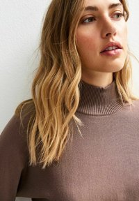 Eine Frau mit langen, welligen blonden Haaren, die einen braunen gerippten Rollkragenpullover trägt und leicht zur Seite schaut, vor einem schlichten Hintergrund.