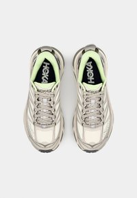 Beige athletic shoes with a textured upper, silver accents, and a light green lining. Features a padded tongue and laces for secure fit.