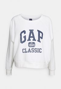 Un sweat-shirt blanc à coupe décontractée, présentant le texte "GAP CLASSIC" en bleu marine et l'année de création, avec un col rond et des poignets côtelés.