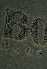 Olivově zelená tkanina s měkkým, vystouplým písmem. Obsahuje "B" a "HUGO" ve strukturovaném designu, což zvýrazňuje povrchové detaily.