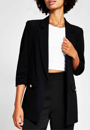 Kvinde iført sort blazer med guldknapper, hvid kort top, sorte bukser og guldkæde, mod en ensartet lys baggrund.