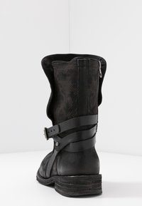 Botte noire en cuir jusqu'au genou avec une finition texturée, motif embossé, fermeture éclair à l'arrière et doubles sangles en cuir autour de la cheville.