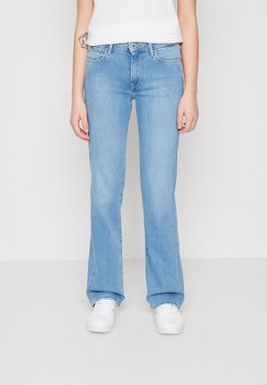Personne portant un jean droit bleu clair et des baskets blanches, debout sur un fond blanc uni.