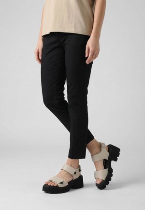 Person trägt schwarze enge Hosen und beige klobige Sandalen mit dicken schwarzen Sohlen, steht vor einem schlichten hellen Hintergrund.