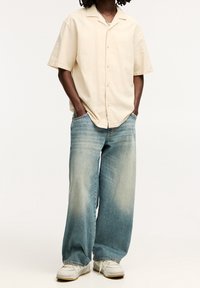 Ljusbeige kortärmad skjorta med knappar, matchad med lösa ljusblå jeans i denim med en gradienttvätt och vita sneakers.