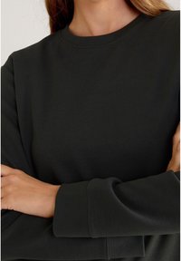 Tmavozelená mikina z měkkého materiálu, s kulatým výstřihem a dlouhými rukávy s žebrovanými manžetami. Detailní pohled na texturu.