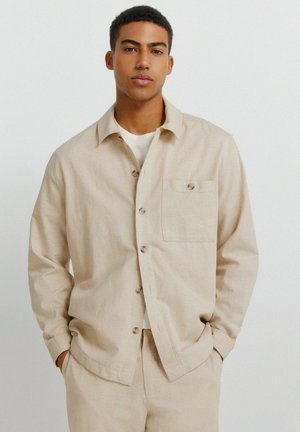 Młody mężczyzna ubrany w beżową koszulę na guziki i dopasowane spodnie, stojący z rękami w kieszeniach na tle jednolitego jasnego tła.