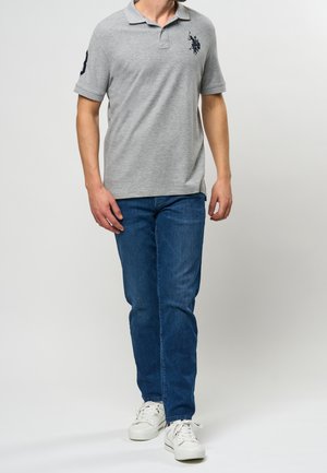 Polo gris de manga corta con un emblema bordado en el pecho izquierdo, combinado con jeans azules y zapatillas blancas.