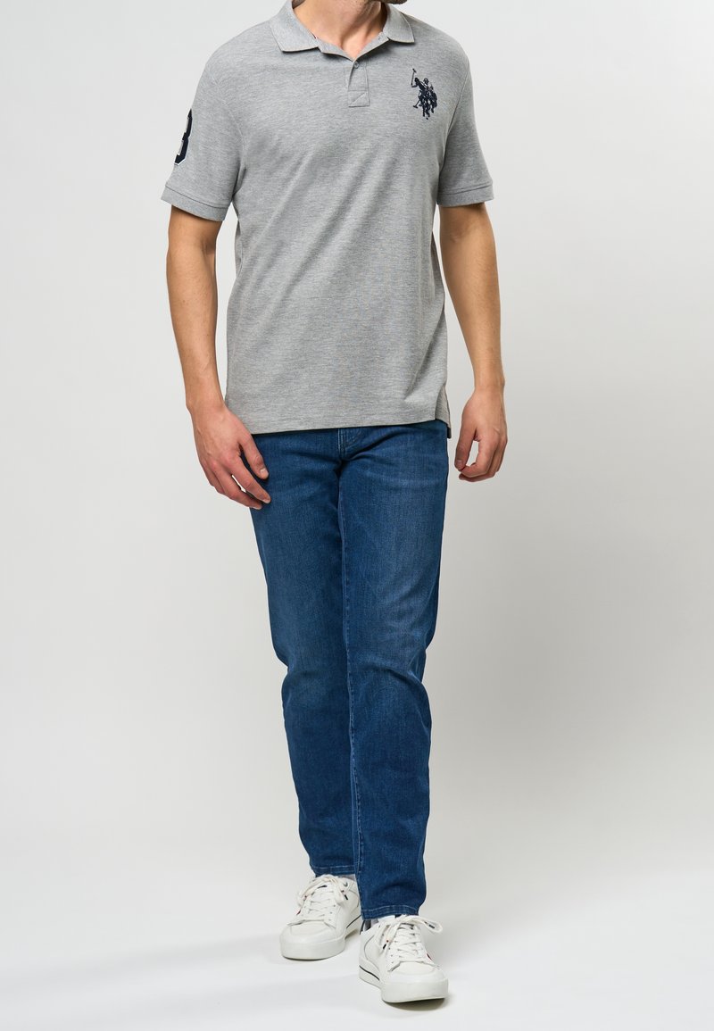 Polo gris à manches courtes avec un emblème brodé sur la poitrine gauche, assorti à un jean bleu et des baskets blanches.