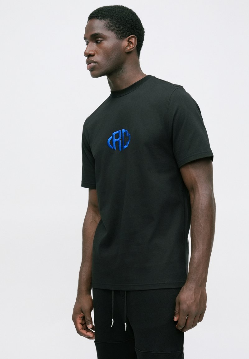 Junger Mann mit schwarzem T-Shirt mit blauem "RC"-Logo, steht seitlich vor einem schlichten weißen Hintergrund.