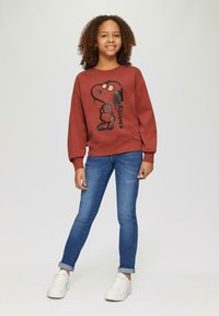 Koraalrood sweatshirt met zwarte Snoopy-print, gecombineerd met een blauwe spijkerbroek en witte sneakers. Zacht materiaal, casual pasvorm, geribbelde boorden.