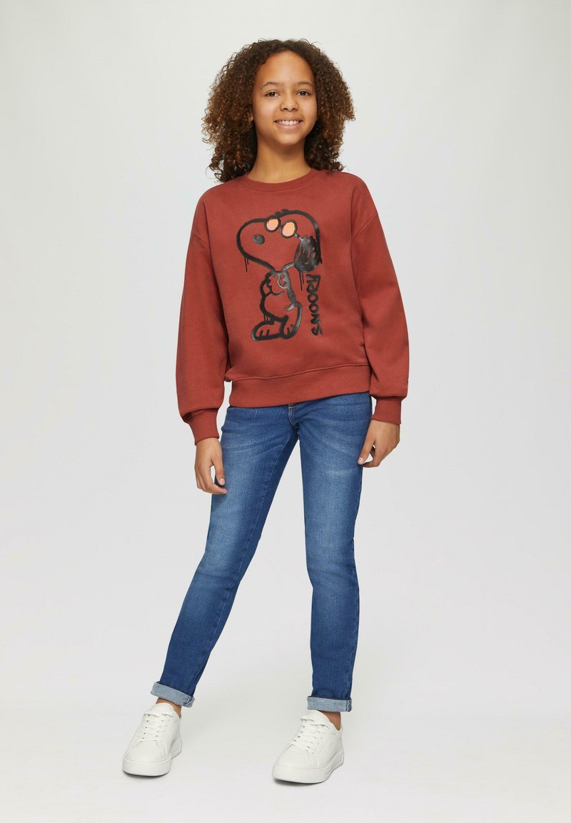 Koraalrood sweatshirt met zwarte Snoopy-print, gecombineerd met een blauwe spijkerbroek en witte sneakers. Zacht materiaal, casual pasvorm, geribbelde boorden.