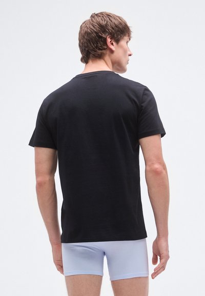 T-shirt preta de algodão, manga curta, corte standard, textura lisa; usada com cuecas boxer azul claro, vista traseira.
